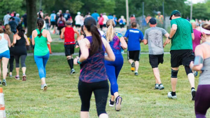 Group of people running outdoors in a 5K race, wearing athletic clothes in various colors, on a grassy course surrounded by trees.