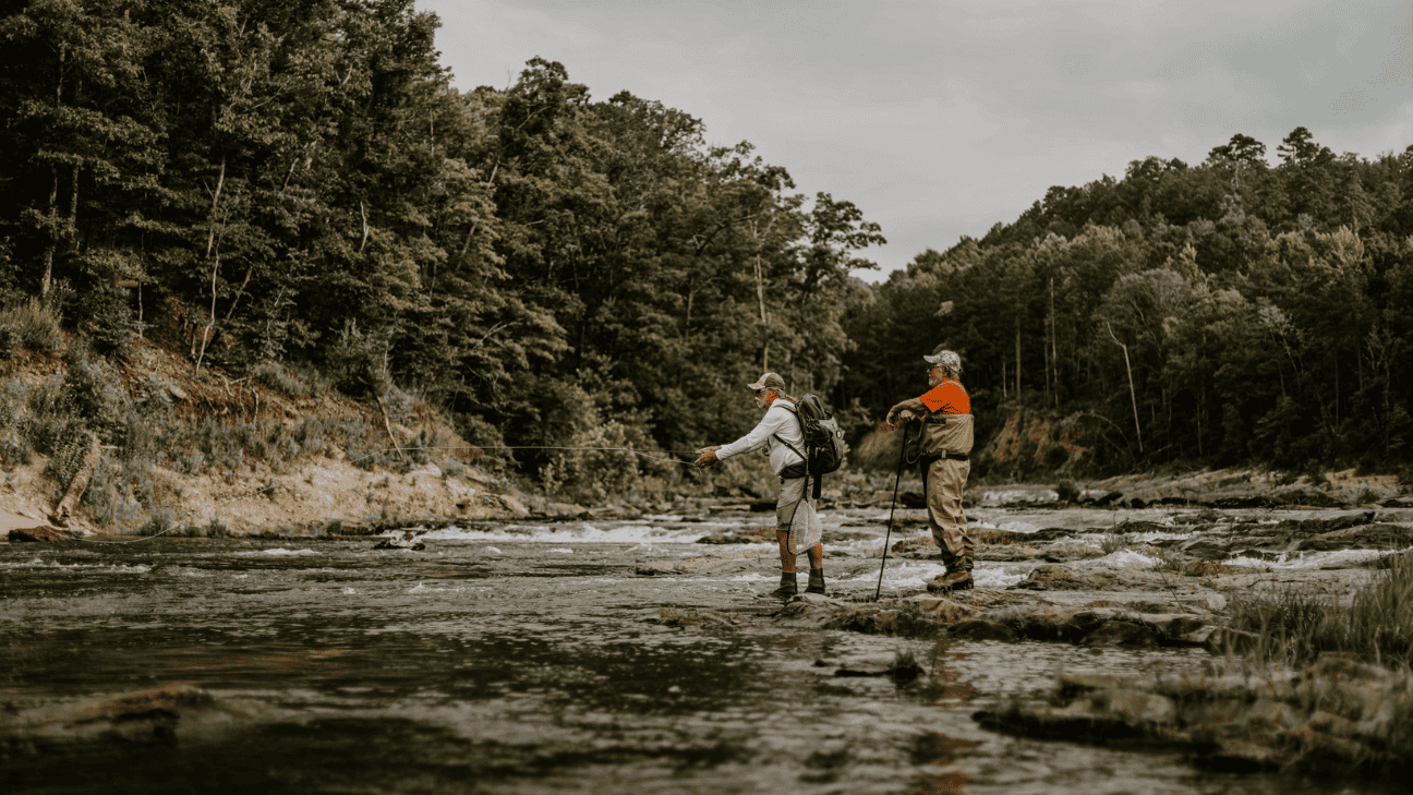Two men fly fishing with one casting a line and the other standing nearby in waders.